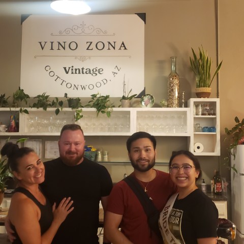 Four people smiling in front of a wall with 'Vino Zona' sign and plants.
