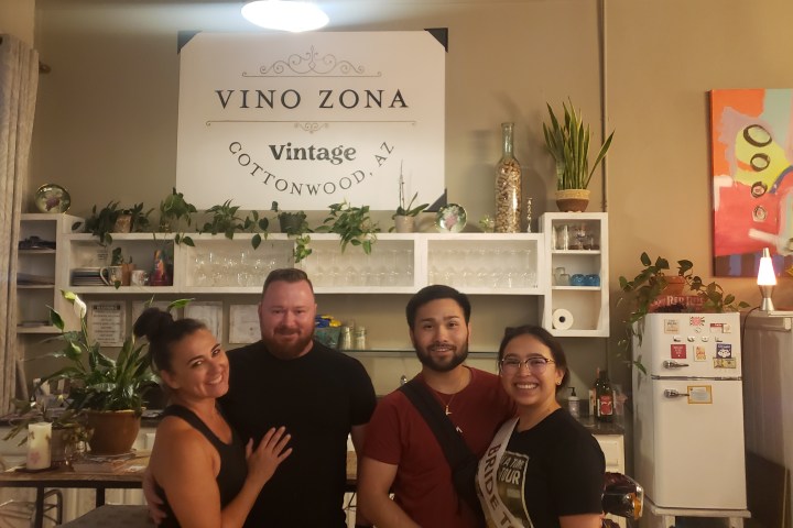 Four people smiling in front of a wall with 'Vino Zona' sign and plants.