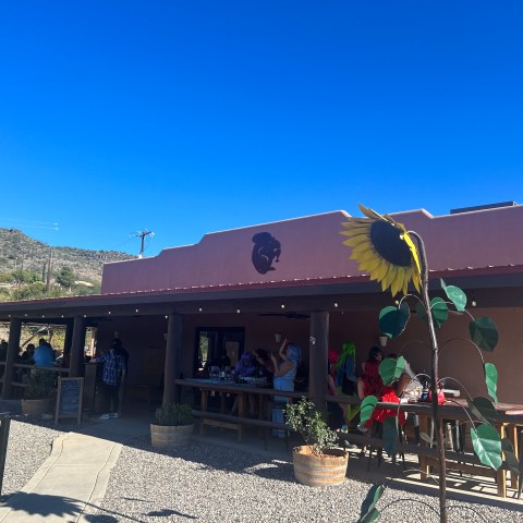 Outdoor cafe with people sitting under a covered patio and a large sunflower sculpture in front.