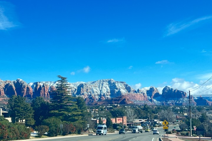 Road view with snow-capped red rocky mountains and clear blue sky in the background.