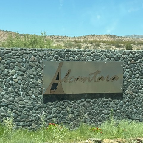 Stone wall with metallic sign reading 'Alcantara' in desert landscape under blue sky.