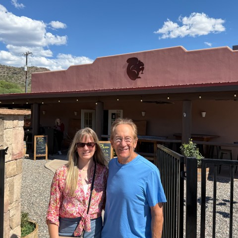 Two people smiling in front of a brown building with a patio and a decorative fence.