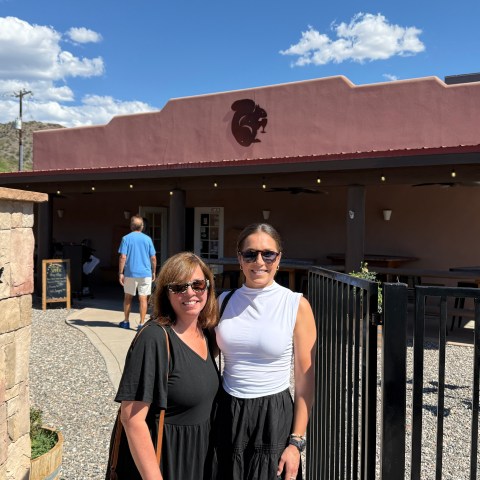 Two people standing in front of a building with a red facade and patio, under a clear blue sky.