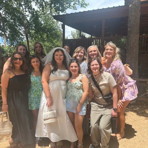 Group of women posing outdoors in summer attire, one in a white dress with 'Bride' bag.
