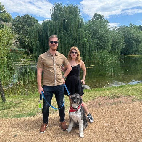 Two people and a dog in front of a pond with lush greenery.