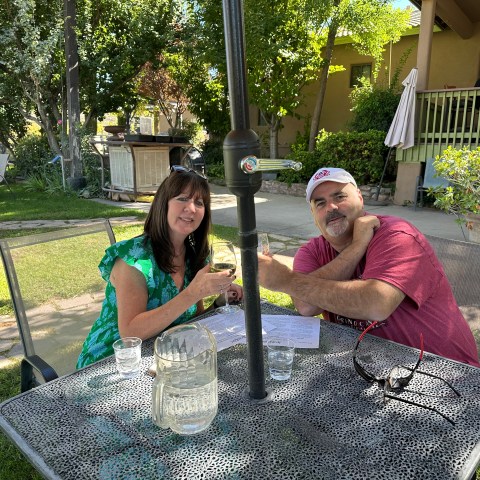 Two people seated at an outdoor table, enjoying drinks in a garden setting with trees and greenery around.