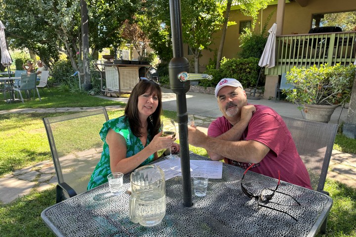 Two people seated at an outdoor table, enjoying drinks in a garden setting with trees and greenery around.