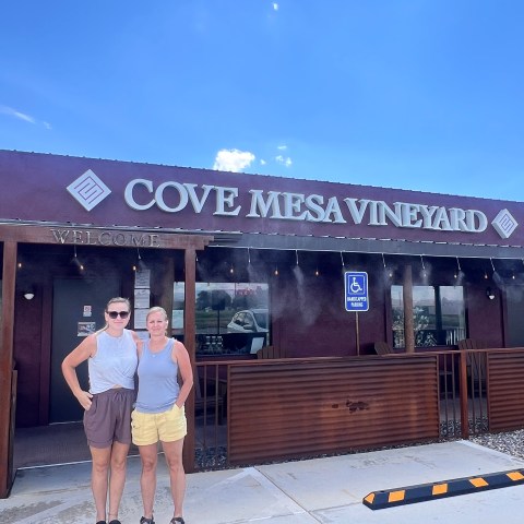 Two people standing in front of Cove Mesa Vineyard building on a sunny day.