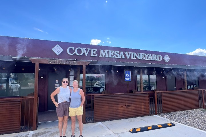 Two people standing in front of Cove Mesa Vineyard building on a sunny day.