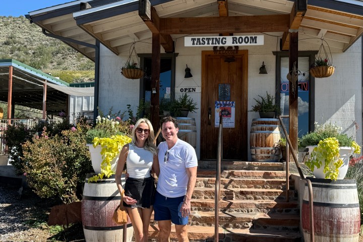 Two people stand in front of a rustic tasting room with plants and barrels.