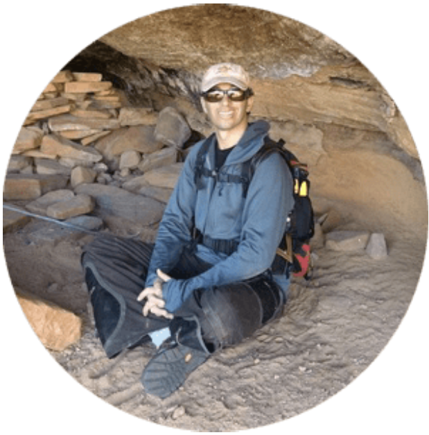 Person sitting outside a rocky cave with backpack and sunglasses.